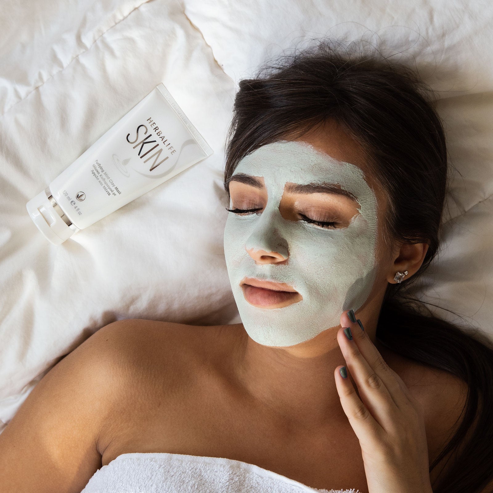 Woman applying a face mask with a product labeled 'Purely for Skin' on a white pillow.
