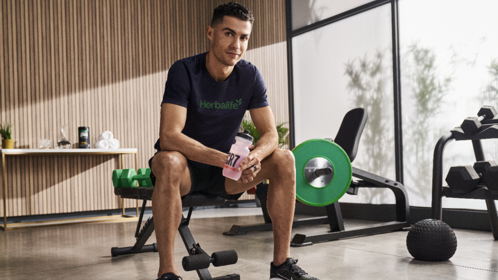 Man sitting on a bench in a gym holding a pink water bottle with a green logo.