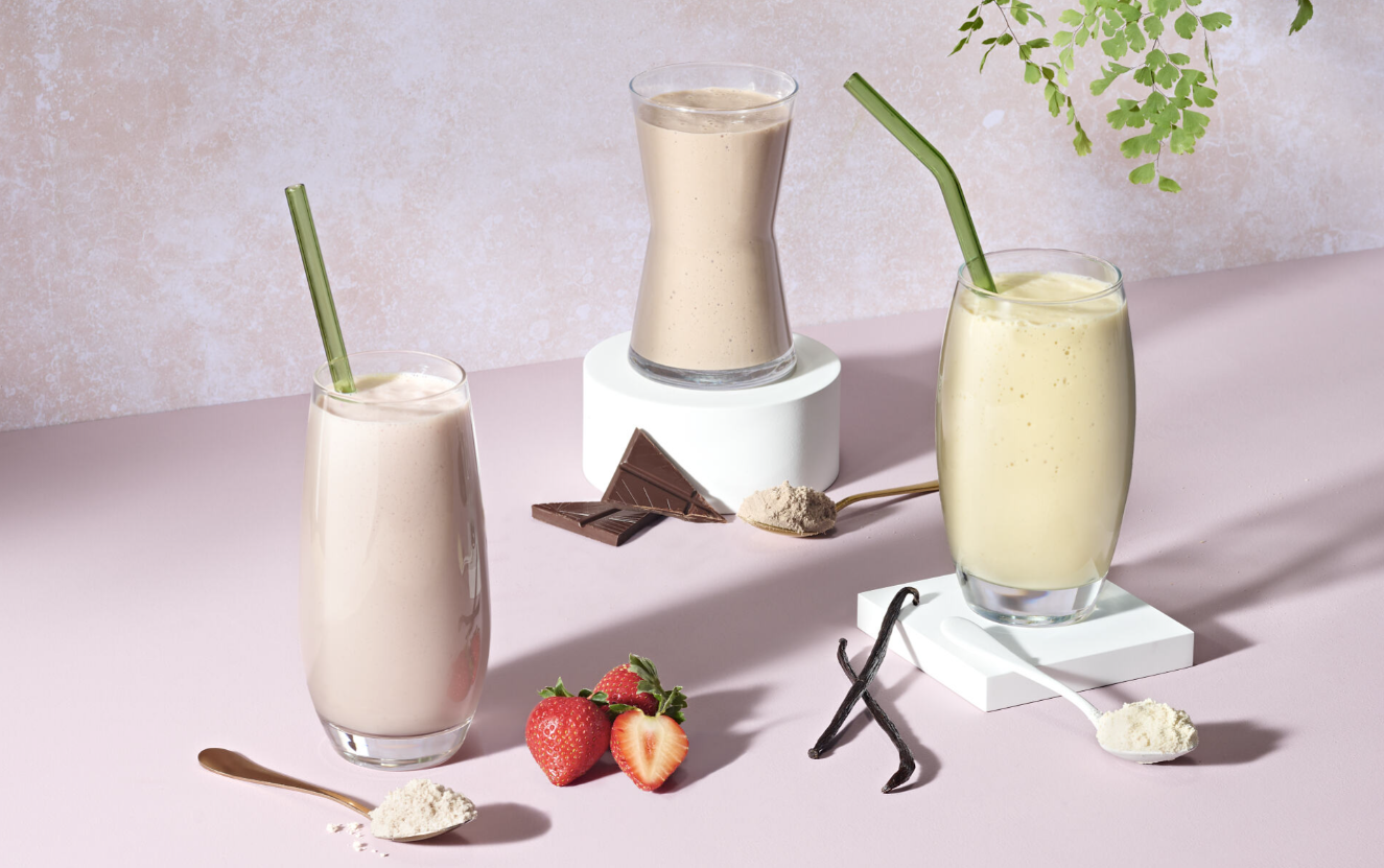 Three glasses of smoothies with straws on a light pink surface, surrounded by strawberries and vanilla beans.
